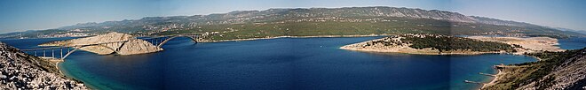 Un bras de mer entre des îles rocheuses ; le pont visible sur la gauche comporte deux arches avec une petite île au milieu.