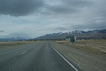 Panneau d'arrivée à Koch-Agatch sur la R256. En arrière-plan, le village se distingue, tandis que des cimes enneigées sont en arrière-plan. Le village se situe dans une steppe plate, celle de la Tchouïa.