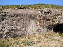 Photographie en couleur montrant un blockhaus en béton armé sur lequel deux traces d'explosions blanches sont visibles, ainsi que des impacts de balles. Le toit du blockhaus est recouvert d'herbe.