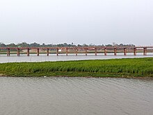 A reddish metal bridge supported by a row of narrow columns crosses horizontally over a stretch of river water, with a grass-covered mudbar in the foreground