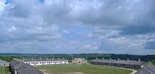 A photograph of a set of long, single-story buildings arranged in a circle around a small grassy field.