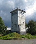 A squat, square white tower on a sloping meadow. Each side of the tower has an unbroken row of windows near the top. There is a searchlight on the tower's flat roof, poles carrying equipment and a railing around the roof's outer edge.