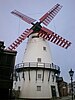 Photograph showing the restored Marsh Mill, a white painted tower windmill with black sails, eleven visible windows and an encircling black wooden stage at the second floor supported by stone corbels