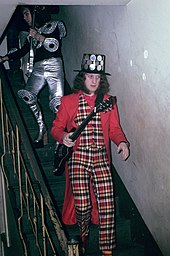 Two men walking down some stairs. In front, the man is wearing a suit, and a jacket on top. He carries a guitar. His hair comes down to his shoulders, and he has large sideburns. On his head is a top hat, covered with large coins. The man following him is wearing metallic plates on his knees, arms and shoulders, and is wearing platform shoes. He carries a guitar, and on his arms is some jewellery. He is wearing a hood of some sort on his head.