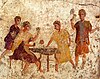An ancient mosaic showing two men playing a game on a small table and two other people standing nearby.