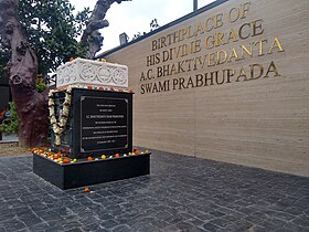 A black rectangular pedestal with an inscription, topped by a white ornate marble slab draped with a flower garland, standing under a double-branched tree.