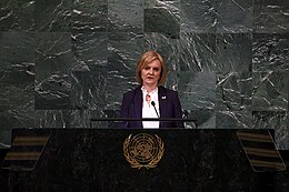 Truss, wearing a white shirt and purple coat, speaks at a podium in front of a greyish-green marble wall.