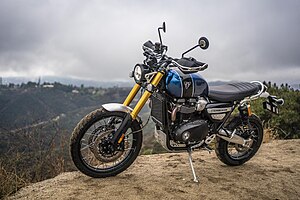 Motorcycle with a blue and black tank, long gold front suspension, and black seat. The motorcycle is parked on its side stand on a dirt road in the mountains, there are houses in the distance.