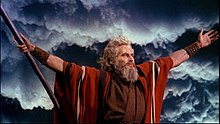 Actor playing Moses wearing a red robe and holding his arms out to the sides with dark clouds behind him
