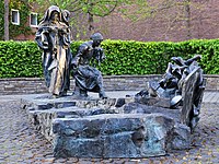 Das Edith-Stein-Denkmal, einer Bronze mit den drei Figuren links und einer nach rechts führenden Rampe, die vor aufgehäuften Schuhen endet. Im Hintergrund eine leuchtend grüne Hecke