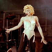 A blond woman onstage with curly hair, and wearing a white corset and black pants. The background is black and smoky.