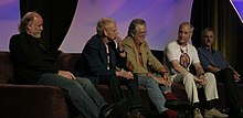Five older men sit on a couch, with an arched, decorative purple background behind them. Ralston, leaning back in his seat, has short hair and wears a shirt with slacks.