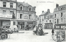 Carte poste en noir et blanc présentant la place Henri IV au début du XXe siècle : des marchandes des quatre-saisons devant la statue, les raisons sociales des magasins peintes sur les murs, un timbre oblitéré en bas à droite.