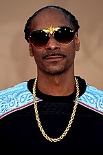 A black man in a gold chain and dark sunglasses stares at the camera