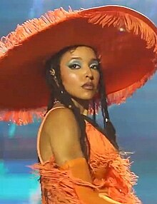 A woman wearing a showy orange costume and big hat dances.