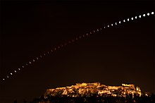 La Lune traverse l'image, passant d'une couleur grise à rouge puis grise. L'acropole est visible éclairée en bas.