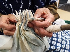 A portion of white fabric with colour-gradient blocks of woven blue pinstripes being tied into cones in the kumo shibori (spider shibori) style by a craftsperson.