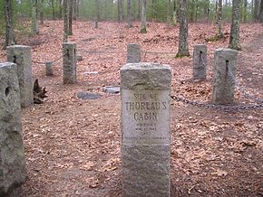 Lieu où Thoreau édifia sa hutte et délimité par des stèles en pierre. Le sol est recouvert de feuilles d'automne. Au fond, à l'arrière-plan : les bois de Walden. Une stèle affichant « Site of Thoreau's cabin » est au premier plan.