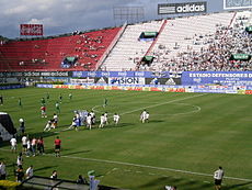 Dois times de futebol, um de verde e outro de branco, preparando-se para iniciar uma partida, em um estádio com arquibancadas vermelhas e brancas, com mais pessoas nesta; o céu está nebuloso e azul.