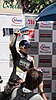 A blond girl with sunglasses in motorcycle racing leathers smiles and holds a silver trophy cup over her head.