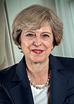 A close-up photograph of Theresa May, a smiling middle-aged woman with short grey hair, a dark blue suit and a necklace in front of light-coloured curtains.