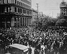 Black and white photo of several hundred people crowding the streets at a downtown intersection
