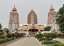 The image showcases the grand entrance to the **Agroha Dham Hindu temple**, with three towering, ornately decorated structures rising prominently against an overcast sky. The central structure is the tallest, with a distinctive brownish-red facade adorned with symmetrically placed windows and carved niches that add depth and texture. Its dome has a rounded, layered design crowned with a golden pinnacle at the top. Flanking the central tower are two slightly shorter towers, each adorned with vibrant colors and intricate artwork. Both towers have sky-blue panels accented with floral patterns and traditional Hindu motifs in hues of pink, gold, and red. The top of each of these side towers is capped with lotus-like structures, topped by golden finials. At the main entrance of the temple, a statue depicting a divine chariot pulled by horses and guided by warriors or deities stands prominently, drawing attention. Below the chariot, a large archway leads into the temple, with detailed architectural elements on either side. The steps leading up to the entrance are wide and surrounded by well-maintained greenery, with various plants, shrubs, and flowering bushes lining the walkway. The scene is bustling with visitors, some ascending the stairs towards the temple, while others are seen standing or moving around in groups. The pathway leading up to the temple is clean and well-kept, bordered by a trimmed lawn and decorative plants, contributing to the peaceful and reverent atmosphere.