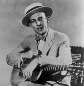 Black and white studio portrait of Jimmie Rodgers sitting on a chair looking at the camera. He wears a tan suit, a white shirt, a bow tie, and a boater hat. He holds a guitar to his right knee with his forearms with his right hand on top of the body and his left hand over the hole