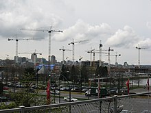 At least a dozen cranes rise over buildings under construction. In the foreground, cars are driving by on existing streets, and a city skyline rises in the background.