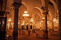 Vue partielle de la salle de prière illuminée, qui montre le mobilier de celle-ci. Outre les lustres et les tapis, ce dernier se compose principalement d'étagères et de bibliothèques.