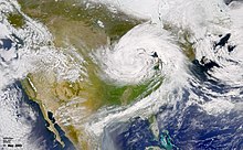 Satellite image of an expansive cloud system over North America characterized by a cyclone near the center of the image and a line of clouds emanating downwards from the cyclone.