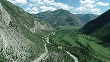 Photographie aérienne d'une vallée encaissée, avec la route longeant la rivière qui passe au centre. L'endroit est vert, avec des arbres recouvrant les pentes. La hauteur des montagnes est importante.