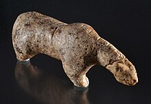 An eggshell coloured, right-facing ivory figure of a four-legged animal with arched shoulders, an elongated low neck and small ears. The surface of the ivory is covered with deep notches and discolouration. The paws are broken off and there is incised damage to the neck.