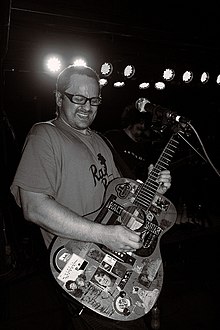 black-and-white image of Brendan B. Brown wearing a print t-shirt and playing an acoustic guitar covered in stickers onstage, in front of a microphone
