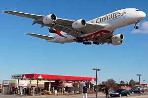 מטוס איירבוס A380 של חברת אמירייטס, שניות ספורות לקראת נחיתה. המטוס, שעל דפנותיו הכתובת "אמירייטס" חולף בגובה נמוך מעל תחנת דלק שעליה השלט "פטרו קנדה".