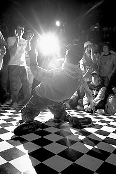 A close-up black and white photo of a male hip-hop dancer surrounded by a small crowd in a nightclub while performing on a checkerboard dance floor.