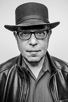 A man with glasses and felt hat wearing a leather jacket.