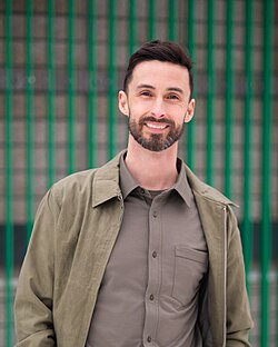 Jordan Wilkie wearing an olive-green shirt and jacket, grinning at camera