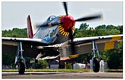 A single nosed-propeller aircraft is on the ground on its wheels with the propeller in motion. The aircraft is viewed from the front, but the red nose faces slightly to the right. The aircraft has black propellers. Parts of the wings and propellers are cropped from view.