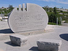 Photo couleur d'une pierre commémorative grise de forme demi-circulaire. Des petits bancs de pierre sont présents devant le mémorial, qui comporte des inscriptions en français et en anglais.