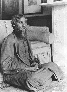 Black-and-white photograph of a bearded middle-aged man dressed in dark robes. He is seated on the floor of an elegantly appointed room and is in front of a plush sofa; he gazes fixedly away to the right, away from the camera.