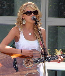 Taylor Swift wearing big sunglasses and playing a wooden acoustic guitar