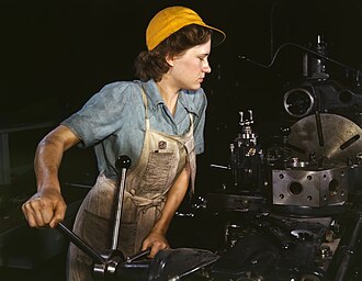 alt= Eine Frau in blauer Arbeitskleidung und mit gelbem Schutzhelm steht an einer großen Industriemaschine und bearbeitet konzentriert ein Flugzeugwerkstück. Im Hintergrund sind weitere Maschinen und Werkzeuge zu sehen.