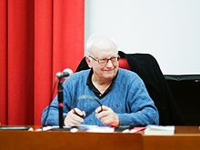 Étienne Balibar in a blue pullover sweater, at a desk with a microphone.