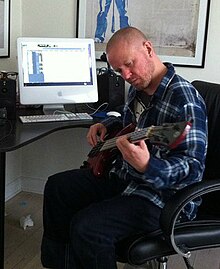 Anders Matthesen sitting in an office chair, playing guitar