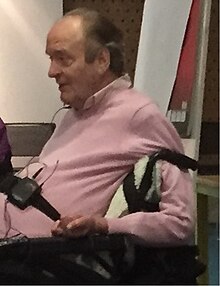 This is a side-on mid-shot of John Evans, sat in his wheelchair, wearing a pink jumper.