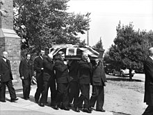 Black and white photo of a coffin which has been draped with a British flag being carried by pallbearers. The pallbearers are middle-aged men wearing formal suits, and three other middle aged men in suits are following the pallbearers.