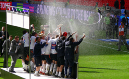 A scene of unmitigated jubilation. Viewed from the right, a crowd of men in a varying assortment of grey suits, navy tracksuits and white, orange and navy soccer attire celebrate together on a podium, many of them brandishing potent bottles of champagne. Some of the men wear straw boaters marked with white, orange and navy ribbons. One of the champagne bottles, which apparently was uncorked rather over-enthusiastically, can be seen in mid-air flying off the front of the podium, its contents flowing forth vigorously.