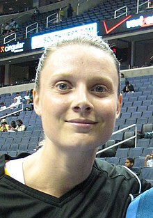 Head of a woman with her hair pulled back tightly wearing a black shirt with arena seats behind her.