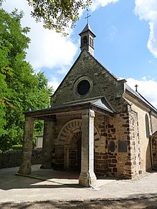 Photographie de la façade d'une chapelle romane. Entrée surmontée d'une voute en pierres alternativement claires et sombres, protégée par un auvent porté par deux colonnes carrées. Un œil de bœuf au-dessus de l'auvent ; un petit clocheton en faîte.